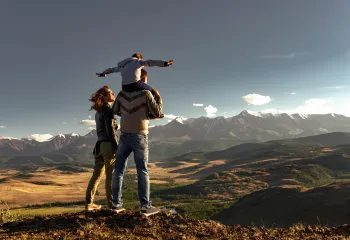 family of three standing on a summit looking down at a valley, mountains in the background